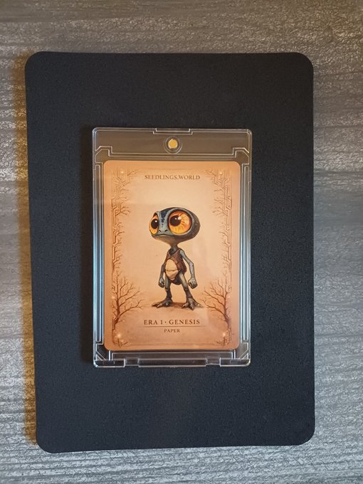 A trading card featuring an illustration of a small alien-like creature with large orange eyes, labeled 'SEEDLING'S WORLD' and 'ERA 1 - GENESIS PAPER', encased in a clear plastic sleeve on a black mat.