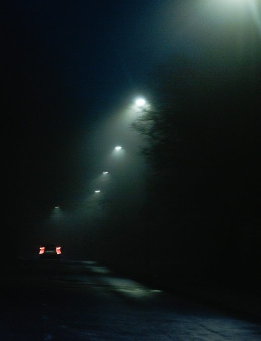 A moody, dark photograph of a foggy road at night illuminated by a row of street lamps receding into the distance, with red taillights of a car visible.
