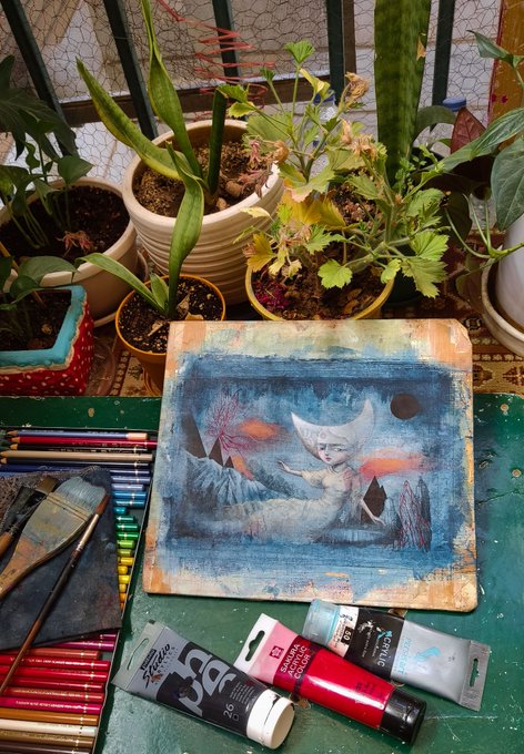 An artistic workspace featuring a watercolor painting of a crescent moon-faced figure on a blue cosmic background, surrounded by potted plants, colored pencils, and tubes of acrylic paint.