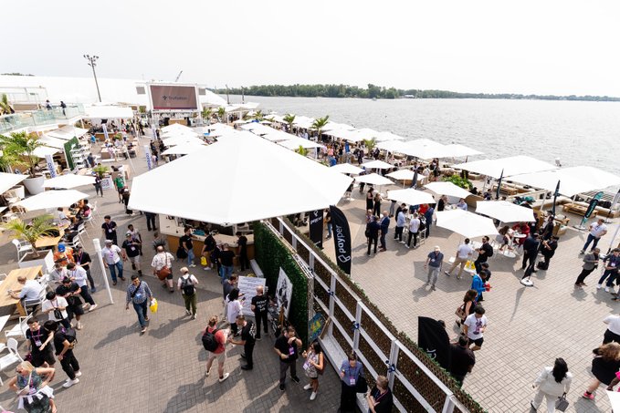 An overhead view captures a bustling waterfront networking event at Consensus, with crowds gathered under white tents and umbrellas beside a lake, reflecting the post's excitement about premier crypto networking opportunities.

Visible text includes "consensus" on the main stage screen and "eToro" branding on a vertical banner.