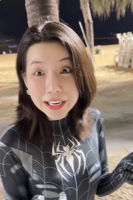 A young woman with shoulder-length hair, wearing a black and white spider-web patterned outfit, smiles at the camera in what appears to be a tropical nighttime setting.