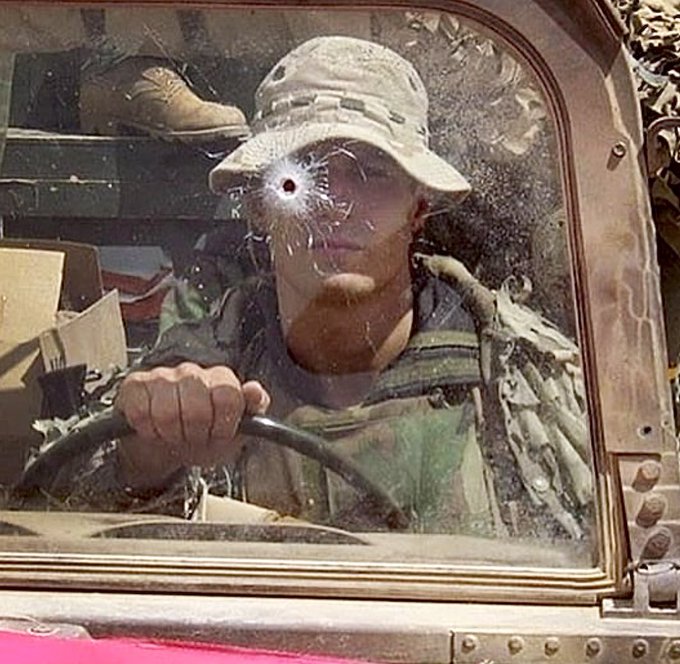 A soldier in camouflage gear and a boonie hat is driving a vehicle with a cracked windshield featuring a bullet hole, reflecting his face.