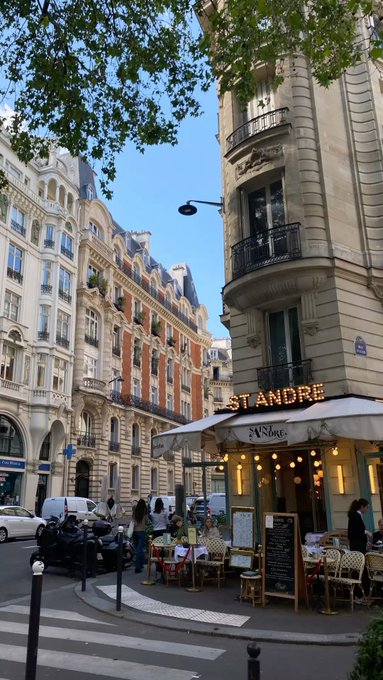 A street corner in Paris with classic Haussmann-style architecture, featuring a cafe named "ST ANDRE" with outdoor seating and a person on a motorcycle passing by.
