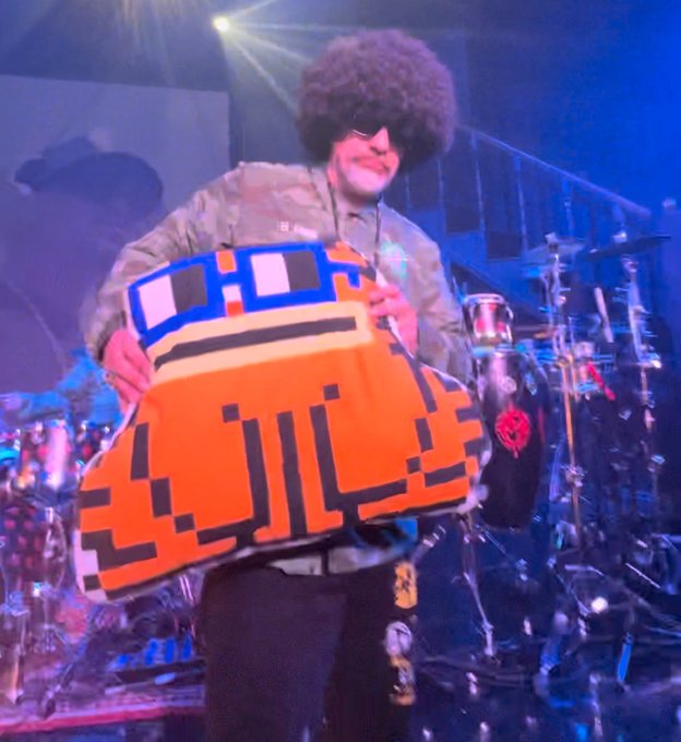 A performer on stage wearing a large afro wig, sunglasses, and a camouflage jacket holds up a pixelated orange and blue torso costume, with a drum set visible in the background.