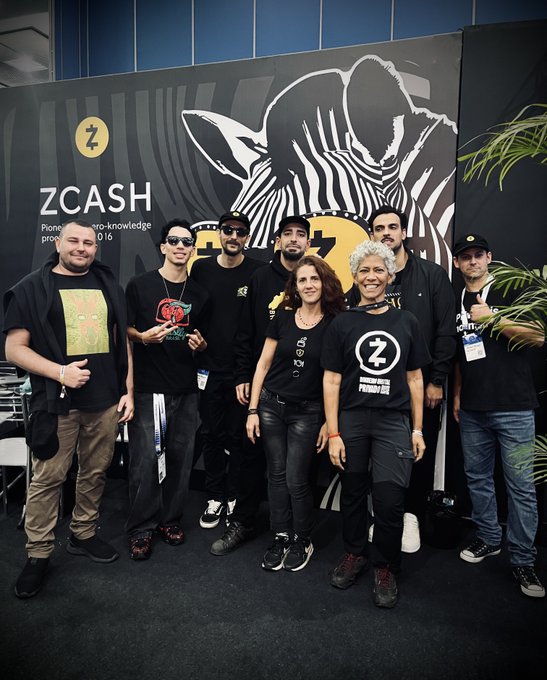 A group of eight people pose in front of a large ZCASH banner featuring a zebra logo and the tagline involving 'zero-knowledge' and '2016.' Several individuals wear branded apparel, including a 'Z' logo shirt, while one person holds a colorful physical Zcash coin.
