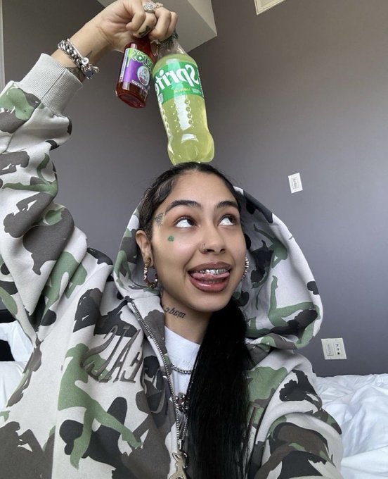 A selfie of a smiling woman in a camouflage hoodie holding a Sprite bottle and a smaller beverage bottle above her head, with visible facial piercings and tattoos.