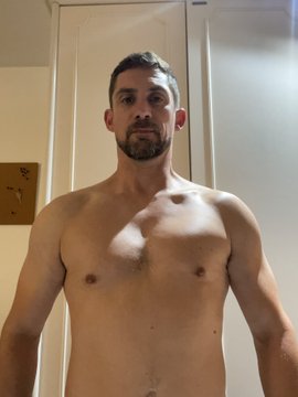 A shirtless man with short hair and a beard stands indoors, posing for a mirror selfie in warm lighting.