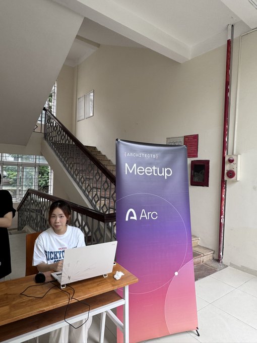 A man sits at a wooden table with a laptop beside an "Arc Meetup" banner, representing Arc Vietnam Campus #2 at Phuong Dong University, an event bringing together builders and IT students to learn about blockchain and Arc.