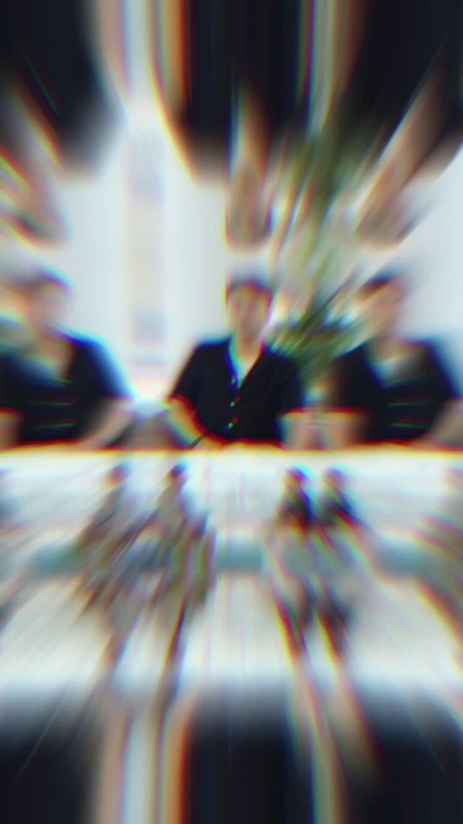 The image shows a blurry photo of people sitting around a table, possibly engaged in conversation or working together. The focus of the image is on one person who appears to be wearing a black shirt. The background features a white wall with a window that allows natural light into the room. There are also some plants visible in the scene.