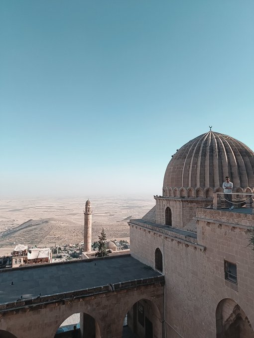 A scenic view from a stone building with a dome overlooking a vast arid landscape, featuring a minaret in the distance and a person standing on a balcony.