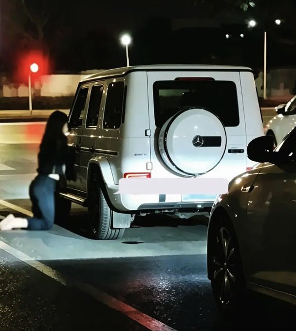 A nighttime scene showing a person kneeling beside a white Mercedes-Benz G-Wagon, with another car visible to the right.
