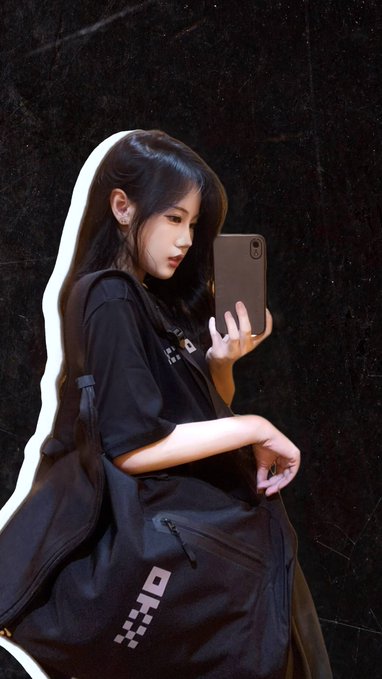 A young woman takes a mirror selfie with her phone while wearing a black hoodie and carrying a black "OKX" branded backpack. The image illustrates her excitement about receiving OKX merchandise, showing off the backpack she plans to use for the gym and the youthful, streetwear-style outfit she loves.