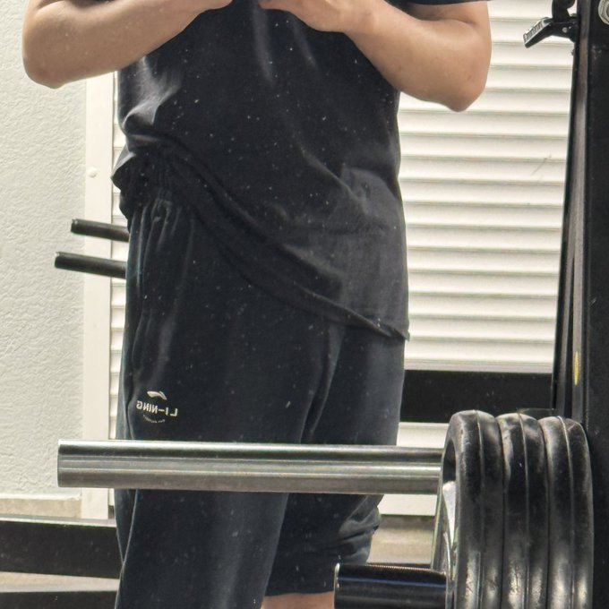 The image shows a person wearing black shorts, standing next to a weight machine at a gym. The person appears to be lifting weights or using the exercise equipment while taking a photo of themselves with their phone. This suggests that they are documenting their fitness journey or sharing their progress on social media platforms.