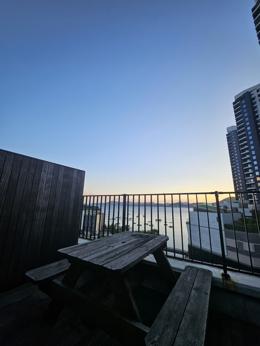 A scenic view from a balcony at dusk, featuring a wooden picnic table in the foreground overlooking a body of water with a city skyline and tall buildings visible in the distance.