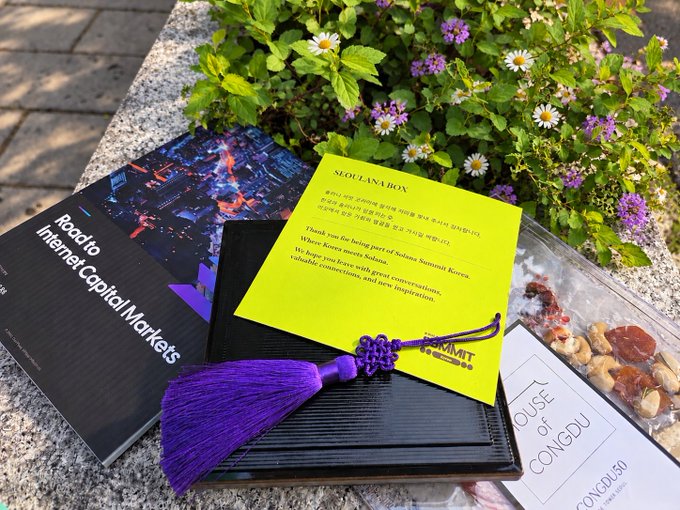 A 'SEOULANABOX' promotional item sits on stone next to flowers, containing a booklet titled 'Road to Internet Capital Markets', a purple tassel bookmark, a 'HOUSE OF CONGDU' card, and dried fruit treats.