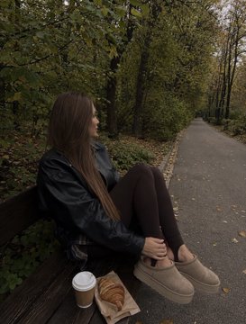 A young woman with long brown hair, wearing a black jacket and tights, sits on a wooden bench along a forest path on a damp autumn day, with a coffee cup and pastry beside her. The cozy outdoor scene matches the post's mention of a rainy autumn day and her IRL busy schedule, while inviting Tezos artists to share their Objkt artworks.
