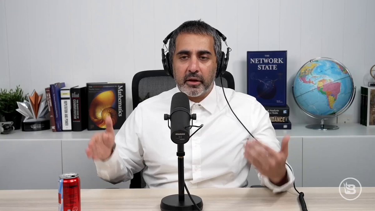 The image features a man wearing headphones, sitting at a desk with a microphone in front of him. The man appears to be talking into the microphone or making gestures while seated. There are several books scattered around the room, suggesting that he might be involved in some form of research or study related to Bitcoin and its impact on America's decline.