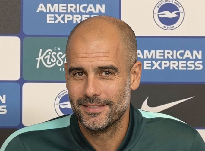 A bald, bearded man wearing a dark green and teal training top is pictured during what appears to be a press conference. Behind him, the backdrop displays sponsor logos including "American Express" and the "Brighton & Hove Albion" crest, along with the Nike swoosh. The setting aligns with the post identifying him as Brighton manager Fabian Hürzeler in 2026.
