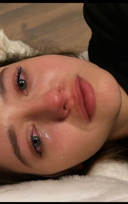 A close-up photo of a woman's face shows her lying down with dewy, glossy skin, dramatic eyelash extensions, and a small tear or droplet near her eye.