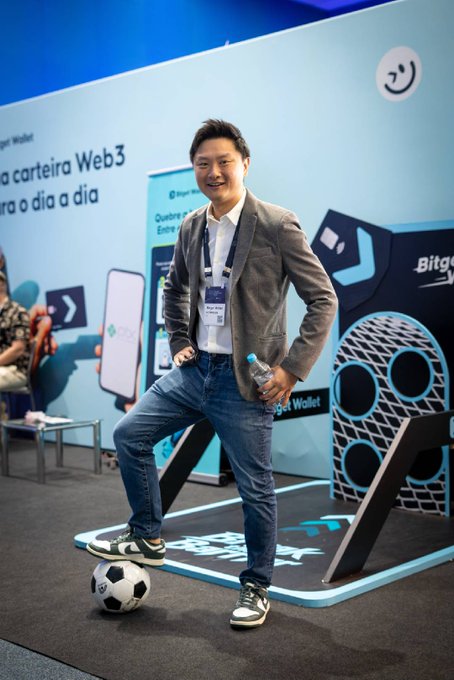 A man in a gray blazer and jeans poses playfully with his foot on a soccer ball at the Bitget Wallet booth (B07) during Blockchain RIO, with visible signage reading "carteira Web3 para o dia a dia" and the Bitget Wallet logo in the background, tying into the post's mention of the crypto payments and trading showcase and the soccer ball giveaway promotion.