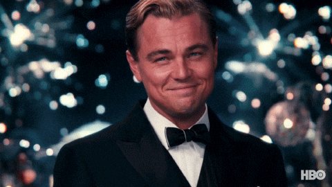 urn of gold and blue fireworks explode against a dark green background, with a man in a black suit and bowtie smiling warmly at the camera.