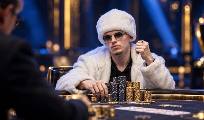 A man wearing a white fur hat, sunglasses, and a matching white fur coat sits at a poker table with stacks of chips.
