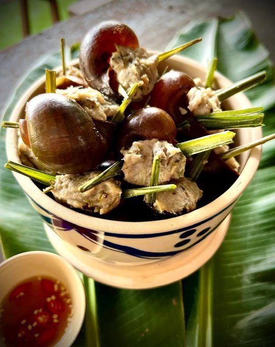 A traditional Vietnamese dish featuring snails cooked with lemongrass stalks served in a decorative ceramic bowl, accompanied by a small dish of red dipping sauce on a banana leaf.