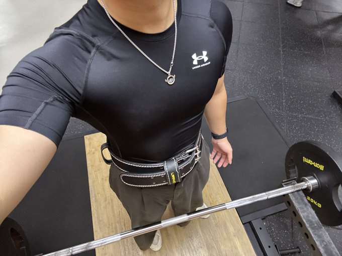 A selfie in a gym showing a person wearing a black Under Armour compression shirt, a silver necklace, and a lifting belt, standing over a loaded barbell on a wooden platform.