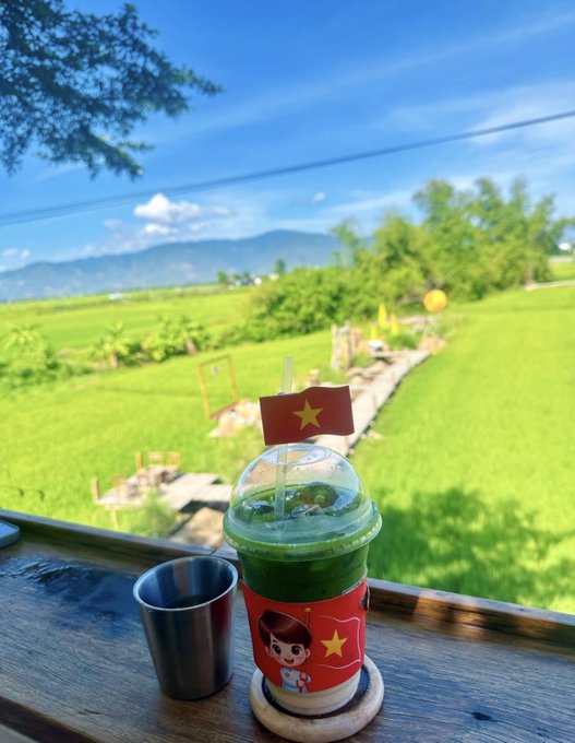 A green smoothie in a clear plastic cup with a small Vietnamese flag and a cartoon character on the sleeve, placed on a wooden ledge overlooking lush green rice fields and mountains.