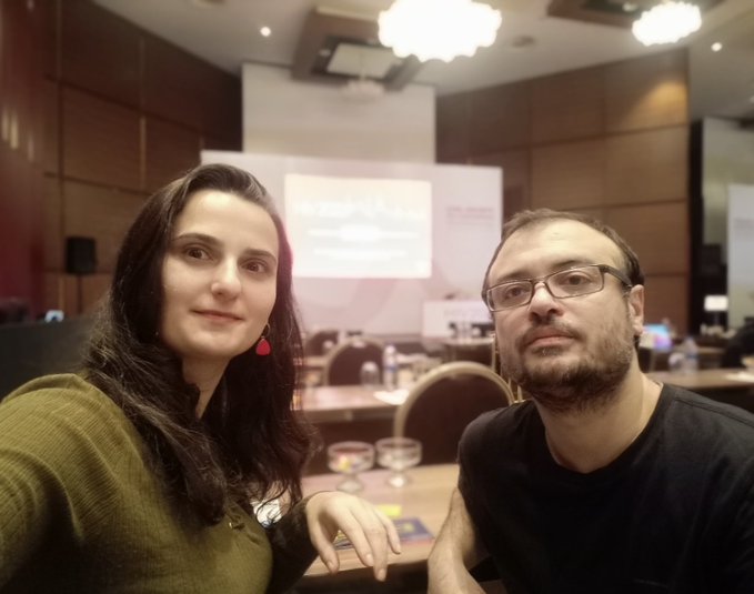 A man and woman take a selfie together at what appears to be a crypto or blockchain industry conference, with a presentation screen visible in the background as they celebrate hitting trading milestones across various platforms like Risextrade, Polymarket, OndoPerps, and Pacifica.