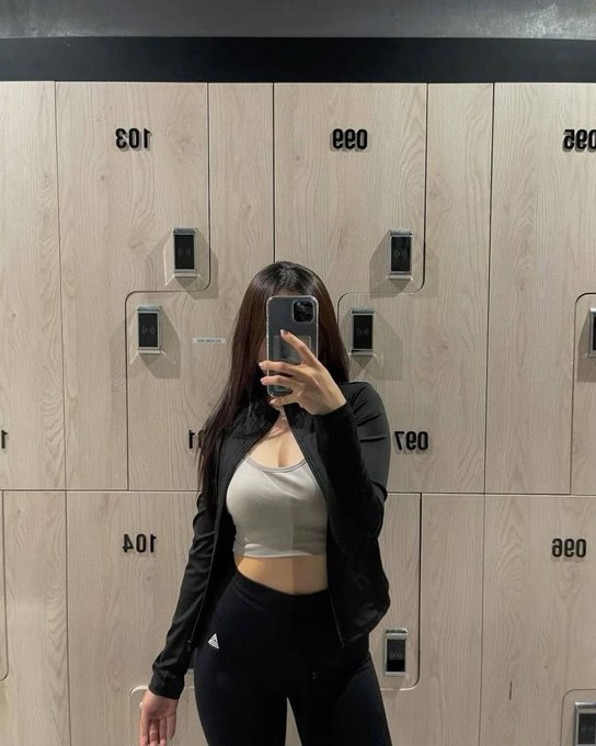 A woman takes a mirror selfie in a locker room, wearing a black Adidas jacket, white crop top, and black leggings, standing in front of lockers labeled '103' and '033'.