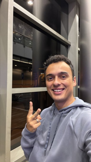 A smiling young man in a gray hoodie poses for a selfie, holding up a peace sign while standing next to a window at night.