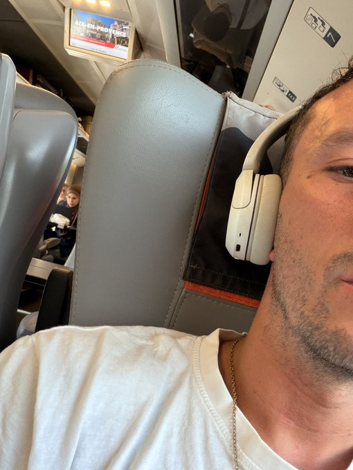 A selfie of a man wearing white AirPods Max headphones and a white t-shirt, sitting in what appears to be a train or airplane with a TV screen visible in the background.