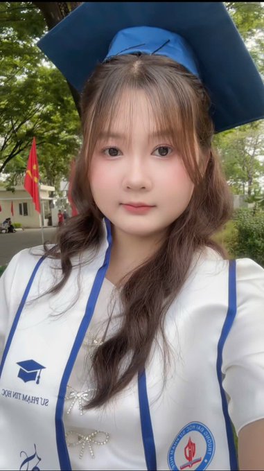 The image features a young woman wearing a blue graduation cap with her hair styled in pigtails. She is dressed in a white shirt that has "our major" written on it, indicating her academic achievement. The background of the photo shows trees, suggesting an outdoor setting or possibly a park where she might have attended her graduation ceremony.