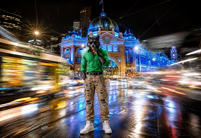 A wolf-headed figure wearing a green shirt and ripped cargo pants stands on a city street at night in front of an illuminated domed building, with motion-blurred traffic passing by.