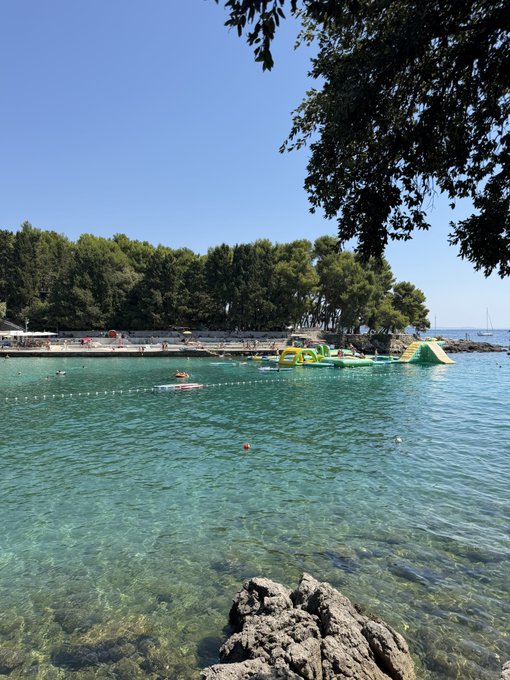 A scenic coastal view featuring clear turquoise water with an inflatable water park floating near a pebble beach, framed by green trees in the foreground and bright blue sky overhead.