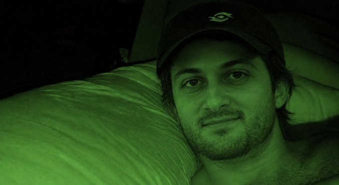 A man wearing a dark baseball cap rests his head on a pillow in a green-tinted night vision style image.