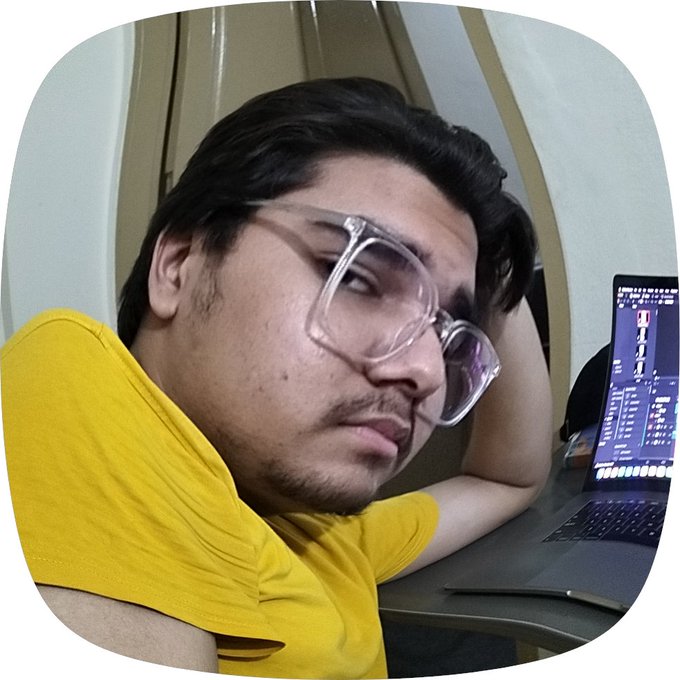 A man wearing clear-framed glasses and a yellow t-shirt takes a selfie next to an open laptop displaying audio editing software.