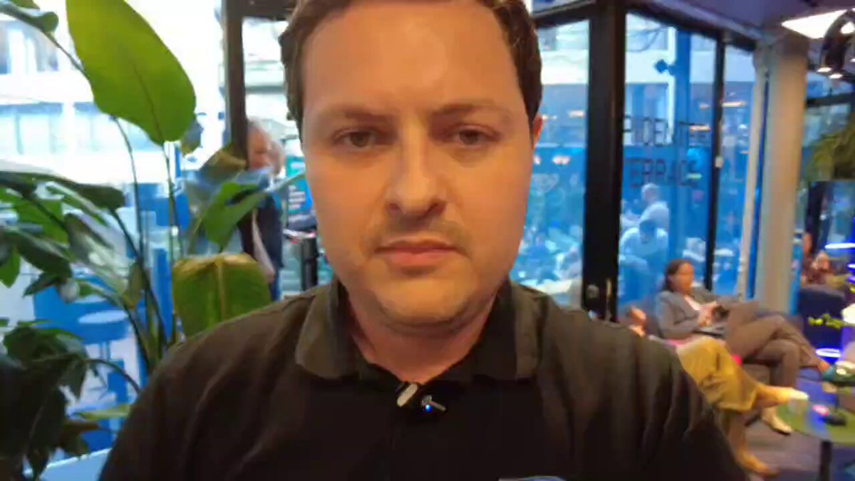 The image shows a man wearing a black shirt with a name tag that reads "Nordic Blockchain Conference". The man appears to be looking directly at the camera, possibly posing for a photo or participating in an online event. In the background, there are other people present who may also be attending the conference. A potted plant can be seen on one side of the room, adding some greenery and life to the space.