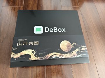 A black DeBox branded box or package on a wooden floor featuring a moon and wave design with Chinese text reading '山月共圆' (Mountain and Moon Together).