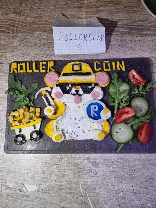 A creative food art display on a black cutting board featuring a pancake decorated to look like a cartoon miner character with 'ROLLER COIN' written in yellow, accompanied by a small mine cart food piece and fresh vegetables, with a handwritten note reading 'ROLLERCOIN' and a heart.
