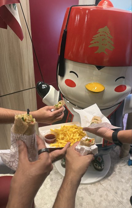 A group of people holding wraps and sharing a basket of french fries with a large red mascot wearing a fez with a Christmas tree on it, inside what appears to be a food court.