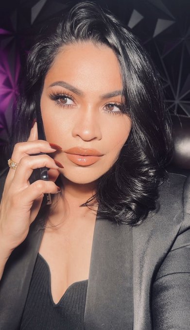 A glamorous portrait of a woman with sleek dark hair, dramatic makeup, and glossy lips, wearing a dark blazer and posing with her hand near her face.