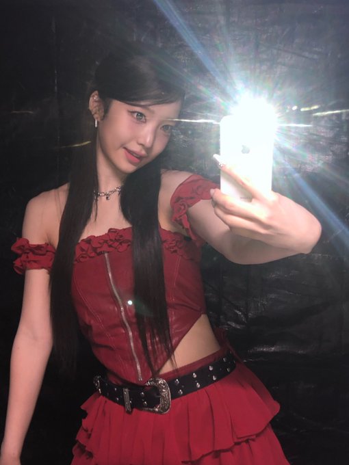 A mirror selfie of a young woman with long dark hair wearing a red corset-style top and skirt with a studded belt, taking the photo with a white iPhone in a dark setting.