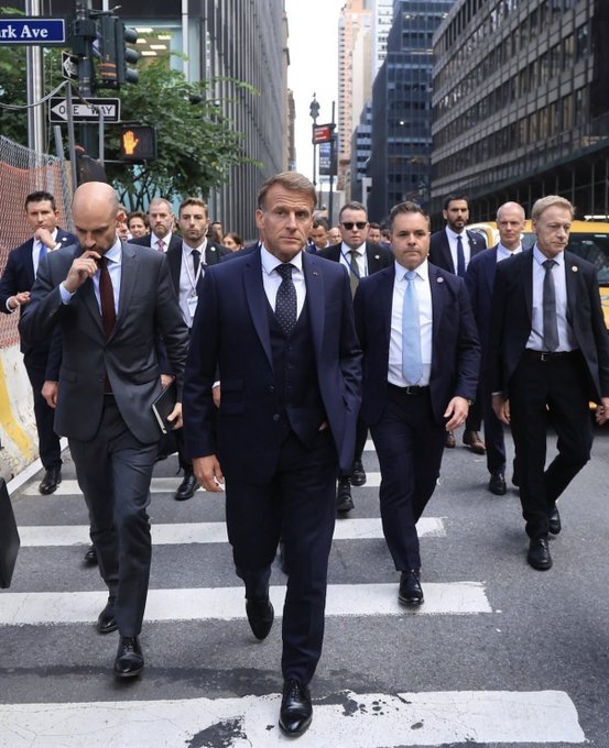 A man in a dark three-piece suit walks across a crosswalk in a city, surrounded by a group of men in suits who appear to be security or staff.