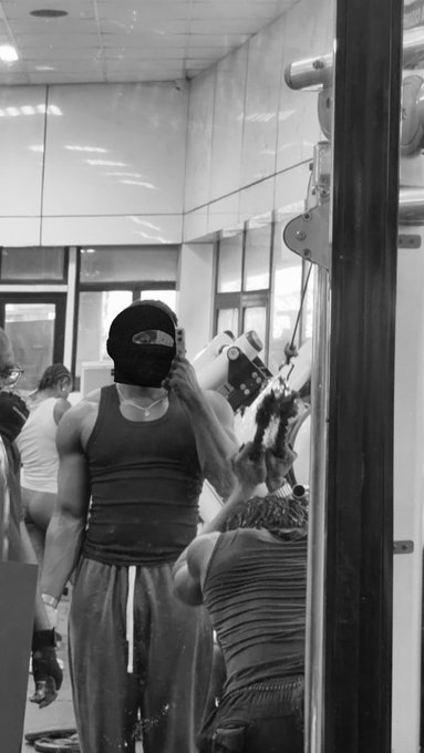 A muscular man in a black tank top and cap holds a shaker bottle in a mirrored gym while flexing toward the camera. The black-and-white photo captures his post-workout pump, aligning with the caption celebrating extra reps inspired by Ansem.