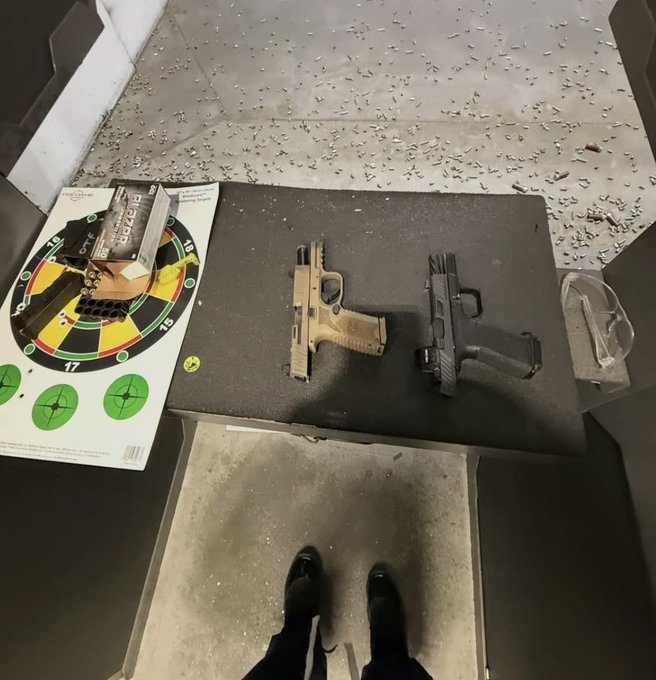 The image shows a pair of feet standing over a gun training table, with two guns resting on top of it - one black and one tan. A target is also visible in the scene. The text "$ALGO Thanks" appears near the bottom right corner of the image.