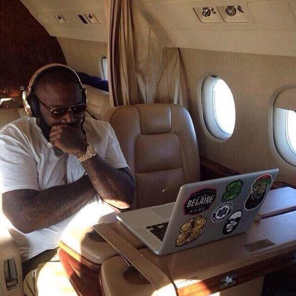 A man wearing headphones and sunglasses sits in a private jet, working on a laptop covered in stickers, including one for 'BELAIRE ROSÉ'.