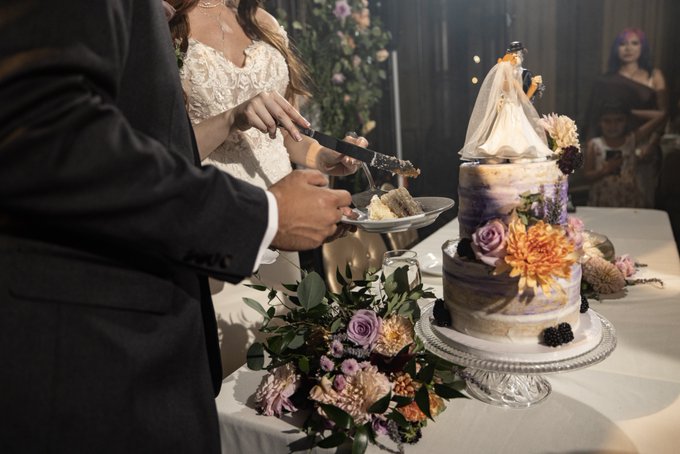 A wedding scene showing a bride and groom cutting a tiered purple and white cake decorated with orange and purple flowers, with a small groom figurine on top.