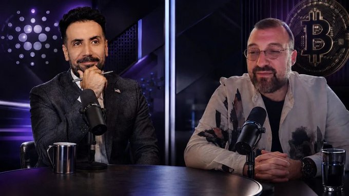 Two men with beards sit at a table with microphones in a studio setting, with a Bitcoin logo visible on the wall behind them.